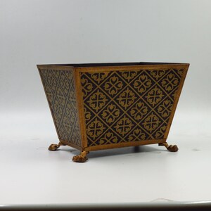 May include: A black metal planter with gold decorative accents. The planter has a square shape with a wide base and a narrow top. The planter has four decorative feet.