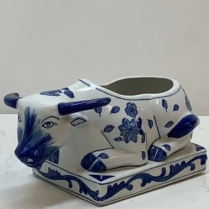 May include: A white ceramic planter shaped like a reclining cow with blue and white floral designs. The planter has a square base with a blue and white floral design.