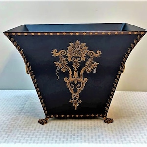May include: A black metal planter with gold accents and a decorative floral design. The planter has a square base and a rounded top with two handles. The planter is sitting on a white surface.