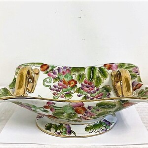 May include: A white porcelain serving bowl with gold handles and a floral design featuring grapes, leaves, and flowers in shades of pink, green, and purple.