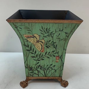 May include: A green metal planter with a butterfly design and gold accents. The planter has three gold feet.