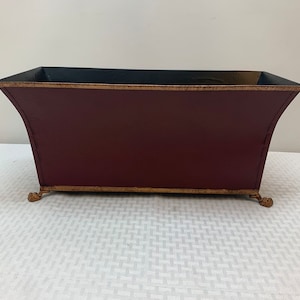 May include: A burgundy metal planter with gold trim and decorative feet. The planter has a black interior.