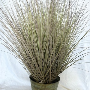 May include: A tall, green artificial grass plant in a weathered metal pot. The plant has long, thin blades of grass that are a mix of light and dark green.