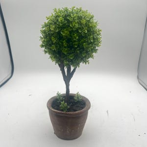 May include: A small, artificial topiary tree with a round, green leafy top. The tree has a brown trunk and is planted in a small, weathered brown pot. The tree is a decorative item.