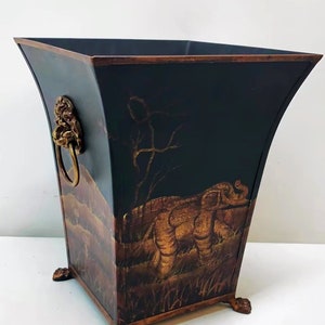 May include: A black metal planter with gold accents and an elephant design. The planter has a square base with four feet and a rounded top. It has a brass handle with a lion head design.