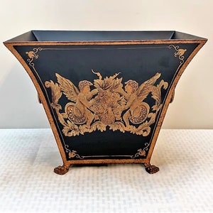 May include: A black metal planter with gold accents and a design of two cherubs with wings and a floral motif. The planter has three gold feet.