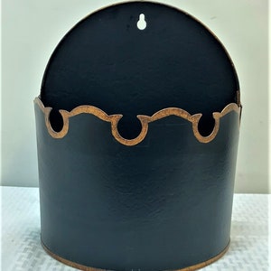May include: Black metal wall mounted planter with a decorative gold trim. The planter has a rounded shape and a small hole at the top for hanging.