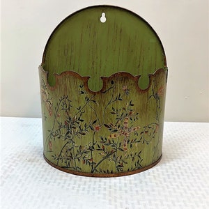 May include: Green metal wall mounted planter with a dragonfly and floral design. The planter has a scalloped top and a hole for hanging.