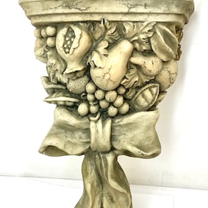 May include: A decorative wall shelf made of plaster or ceramic, featuring a sculpted design of fruit and a bow. The shelf is painted in a light beige color and has a distressed finish.