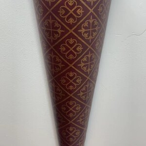 May include: A burgundy metal wall vase with a gold geometric pattern. The vase has a cone shape and a decorative metal finial at the bottom.
