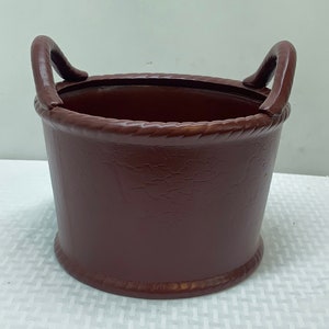 May include: A brown faux leather planter with two handles. The planter has a textured surface and a rope-like trim around the top.