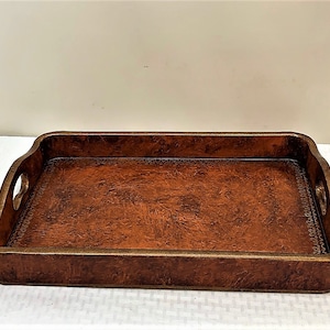 May include: A rectangular brown leather serving tray with a gold rim and two handles on the sides. The tray has a distressed look and a subtle pattern.