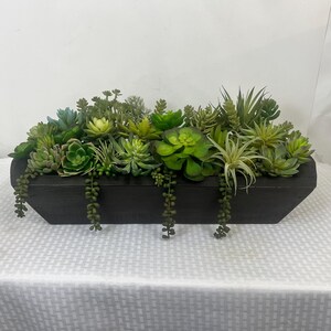 May include: A rectangular wooden planter filled with a variety of green succulents. The planter is dark brown and sits on a white surface. The succulents are various shades of green and have different textures and shapes.