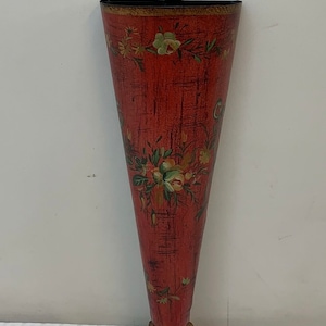 May include: A red metal cone-shaped wall vase with a floral design and a gold-colored base. The vase has a black metal hanger at the top.