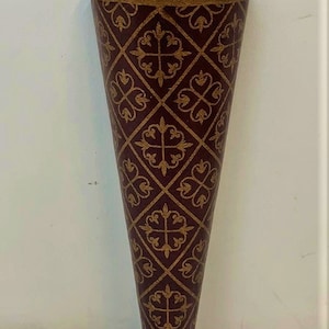 May include: A burgundy and gold metal wall vase with a decorative pattern. The vase has a hanging loop at the top and a decorative base.