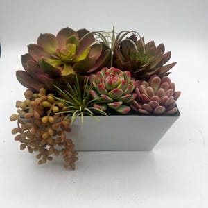 May include: A rectangular white planter filled with a variety of artificial succulents. The succulents are in shades of green, red, and brown. Some have a cascading, trailing habit. The planter has a modern, minimalist design.