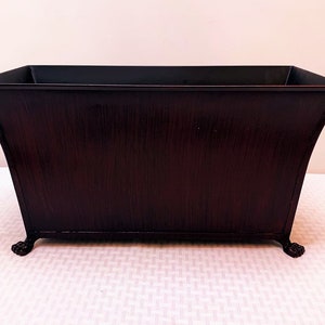 May include: A dark brown metal planter with a rectangular shape and a slightly flared top. The planter has four decorative feet.