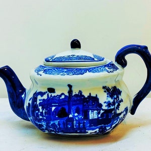 May include: A blue and white porcelain teapot with a floral design and a scene of a building with trees. The teapot has a lid and a handle.