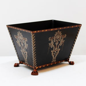 May include: Black metal planter with gold and brown floral designs and copper-colored feet.