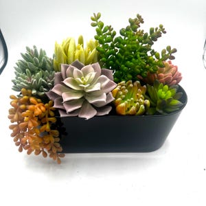 May include: A black rectangular planter filled with a variety of artificial succulents. The succulents are in shades of green, orange, and purple. The arrangement includes a large, light purple succulent in the center.