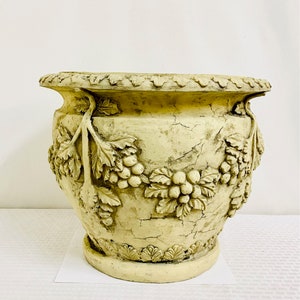 May include: A large, off-white ceramic planter with a distressed finish. The planter is decorated with a grapevine design, featuring leaves and grapes.