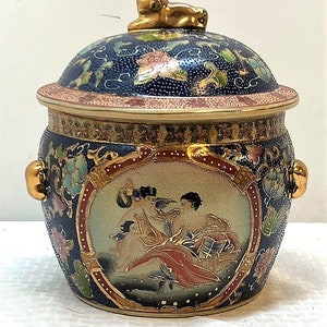 May include: A blue and gold porcelain jar with a lid featuring a floral design and a gold lion finial. The jar has a circular panel with a painted scene of people in traditional clothing.