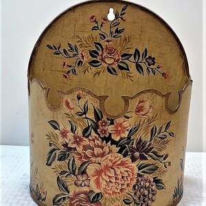 May include: A decorative wall mounted storage container with a scalloped edge and floral pattern. The container is painted in shades of gold, brown, and pink.