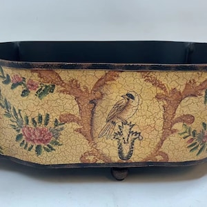 May include: A decorative metal planter with a distressed yellow finish and floral design. The planter has two gold lion head handles and a bird perched on a branch.