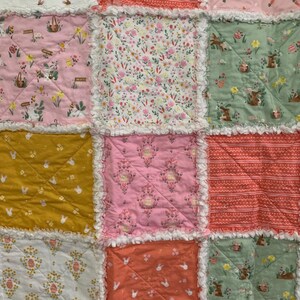 May include: A patchwork quilt with a variety of floral and bunny patterns in pink, green, yellow, and white. The quilt is made with a variety of fabrics and has a ruffled edge.