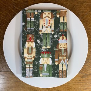 May include: A green fabric napkin with a repeating pattern of nutcracker soldiers. The nutcrackers are wearing colorful uniforms and hats. The background is a dark green with a white snowflake pattern.