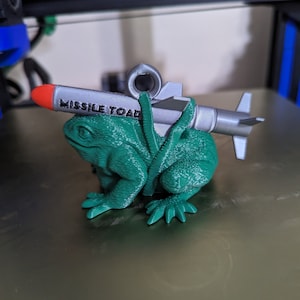 May include: A 3D-printed, green frog figurine with a silver missile attached to its back. The missile has a red tip and the words "MISSILE TOAD" printed on it. The frog is sitting on a reflective surface.