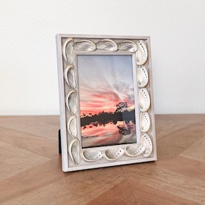 May include: A rectangular photo frame with a light gray wooden border. The frame is decorated with iridescent seashells. The photo inside shows a sunset over a body of water with a tree in the distance. The frame is standing on a wooden surface.
