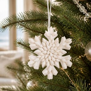 May include: A white snowflake-shaped ornament crafted from small, pointed seashells arranged in a radial pattern. The ornament is suspended by a thin, shimmering silver ribbon and features a textured, multi-layered surface of natural shells.