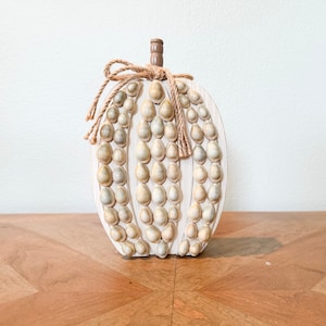 May include: A decorative pumpkin made of wood, painted white, and adorned with small seashells. The pumpkin has a brown stem and a jute twine bow. The design is reminiscent of fall and coastal decor.
