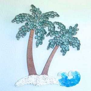 May include: A wall art piece featuring two palm trees with trunks made of brown material and fronds crafted from numerous small, circular, teal-colored shells. The base consists of white crushed shells and a wave made of blue sea glass pieces.