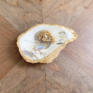 May include: A decorative oyster shell trinket dish with a gold-painted rim and a floral decoupage interior. Resting inside the shell is a delicate gold chain necklace featuring a small, iridescent, oval-shaped pendant with a Kendra Scott tag.