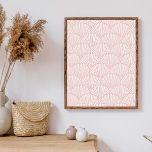 May include: A framed print with a pink and white geometric pattern of overlapping fans. The print is hanging on a white wall above a wooden table with a woven basket, two small vases, and a bouquet of dried pampas grass.