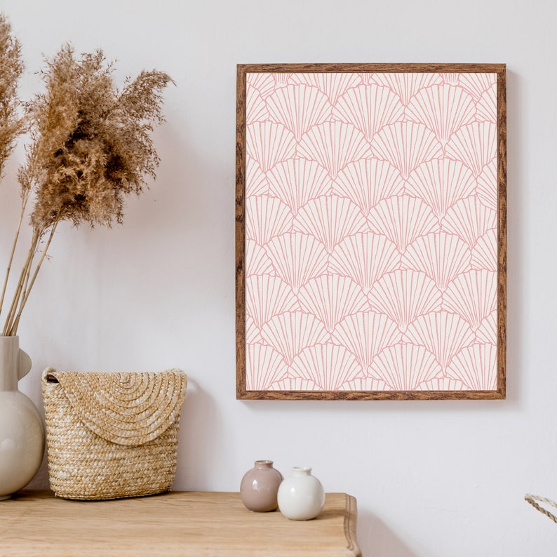 May include: A framed print with a pink and white geometric pattern of overlapping fans. The print is hanging on a white wall above a wooden table with a woven basket, two small vases, and a bouquet of dried pampas grass.