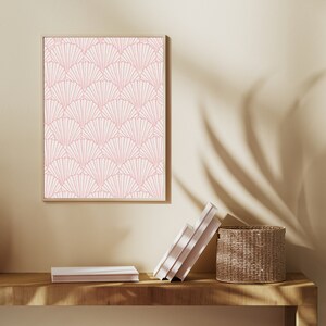 May include: A framed print with a pink and white geometric pattern of overlapping fan shapes. The print is hanging on a beige wall above a wooden shelf with a stack of books and a woven basket.