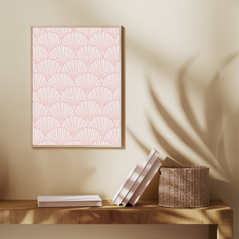 May include: A framed print with a pink and white geometric pattern of overlapping fan shapes. The print is hanging on a beige wall above a wooden shelf with a stack of books and a woven basket.