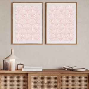 May include: Two framed prints with a pink geometric pattern of overlapping fan shapes on a white background. The prints are hanging on a light beige wall above a wooden cabinet with a woven rattan front.