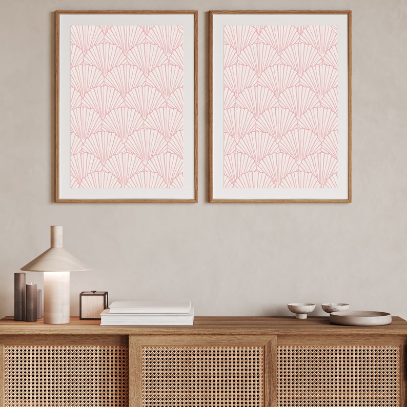 May include: Two framed prints with a pink geometric pattern of overlapping fan shapes on a white background. The prints are hanging on a light beige wall above a wooden cabinet with a woven rattan front.