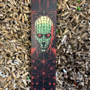 Könnte beinhalten: Ein schwarzes Lesezeichen mit rotem Rand zeigt eine Grafik eines grün-roten Gesichts mit einem Kopf, der mit Nägeln bedeckt ist. Der Hintergrund ist schwarz mit einem rot-weißen geometrischen Muster.