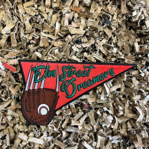 Peut inclure: Un fanion en feutre rouge avec le texte "Elm Street Dreamers" brodé en vert. Un gant de baseball brun avec des coutures blanches et une balle de baseball se trouvent dans le coin supérieur gauche du fanion.