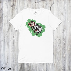 Puede incluir: Camiseta blanca con un gráfico de una vaca en blanco y negro sobre un fondo verde. La vaca tiene una lengua roja y una cara de calavera. El texto "UFO" está impreso en la etiqueta.