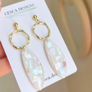 May include: Gold-coloured earrings with a circular top and an elongated, teardrop-shaped bottom. The bottom portion is filled with iridescent, multi-coloured fragments. The earrings are displayed on a white card with the text "CESCA DESIGNS".
