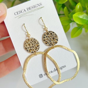 May include: Gold hoop earrings with a dangling leopard print circle. The earrings are on a white card with the text "CESCA DESIGNS" and "@ca.designs".