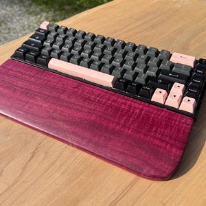May include: A mechanical keyboard with black and pink keycaps, resting on a deep red wooden wrist rest. The keyboard has a compact layout, with a mix of black and pink keys. The wrist rest is made of a richly grained, dark red wood.