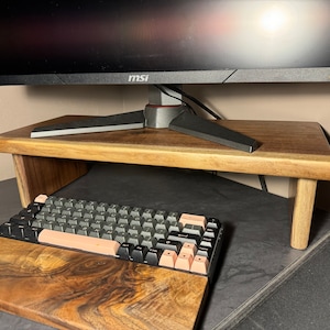May include: A wooden monitor stand with a keyboard and mouse pad on a black surface. The monitor stand is made of dark wood with a natural finish. The keyboard is black with pink keys. The mouse pad is also made of dark wood with a natural finish.