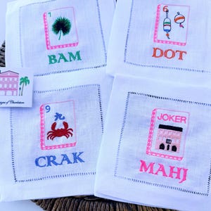 Może przedstawiać: Cztery kwadratowe białe lniane serwetki z wyhaftowanymi wzorami płytek mahjong. Każda serwetka przedstawia unikalną grafikę z napisami BAM, DOT, CRAK i MAHJ, wykonanymi przy użyciu żywych kolor&oacute;w, takich jak zielony, pomarańczowy, niebieski i r&oacute;żowy.